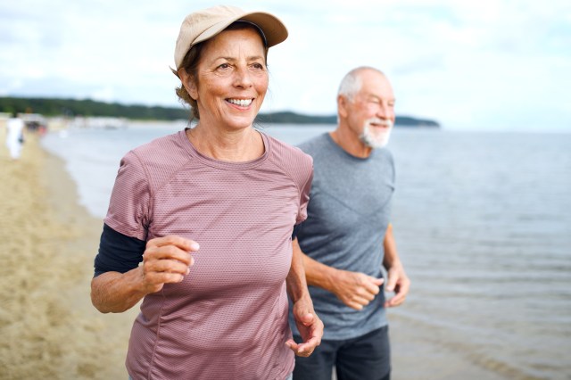 Active senior couple runners jogging outdoors on sandy beach by sea in early morning