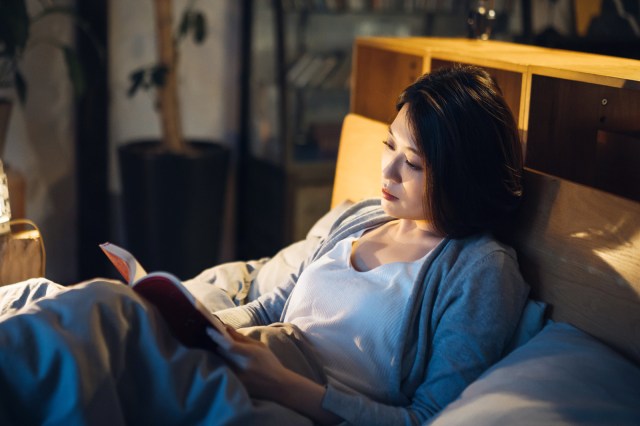 Relaxed young Asian woman lying in bed and reading a book before bedtime at night