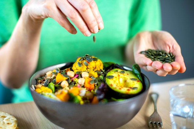 Close Up Of Woman Adding Pumpkin Seeds To Vegan Bowl